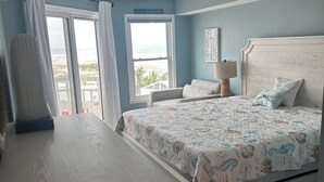Un dormitorio con cama, mesita de noche, lámpara y vistas a la playa a través del ventanal.