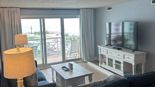 Una sala de estar con un sofá, una mesa de centro, una lámpara, un televisor sobre un mueble blanco y vistas al mar a través de amplios ventanales.