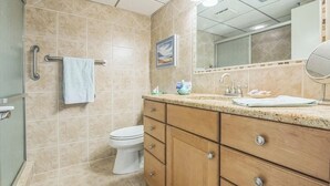 Un baño con baldosas color crema, un inodoro blanco, un tocador de madera con cajones, un perchero para toallas y un espejo.