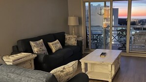 Sala de estar con un sofá oscuro, una mesa de centro blanca y una lámpara. Se ve el exterior a través de un ventanal.