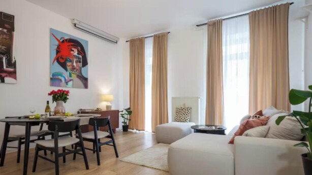 Un salon moderne avec un coin repas, comprenant un canapé, une table basse et une table à manger avec des chaises. De grandes fenêtres sont agrémentées de rideaux, un tableau orne le mur et une plante en pot est posée dans un coin.