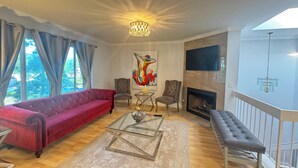 Ein Wohnzimmer mit einem roten Sofa, einem Glastisch, zwei Sesseln, einem Kamin und einem großen Fenster mit Vorhängen.