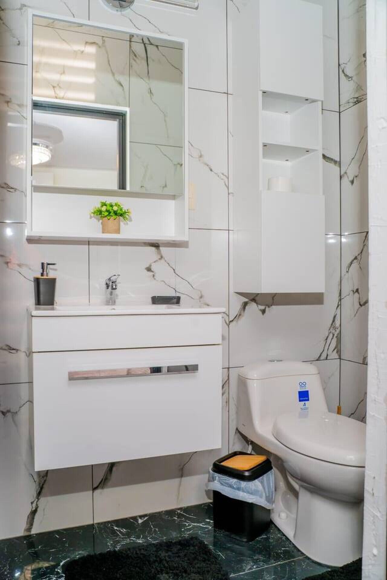 Un baño moderno con detalles de mármol, un tocador blanco con espejo, inodoro y un cubo de basura.