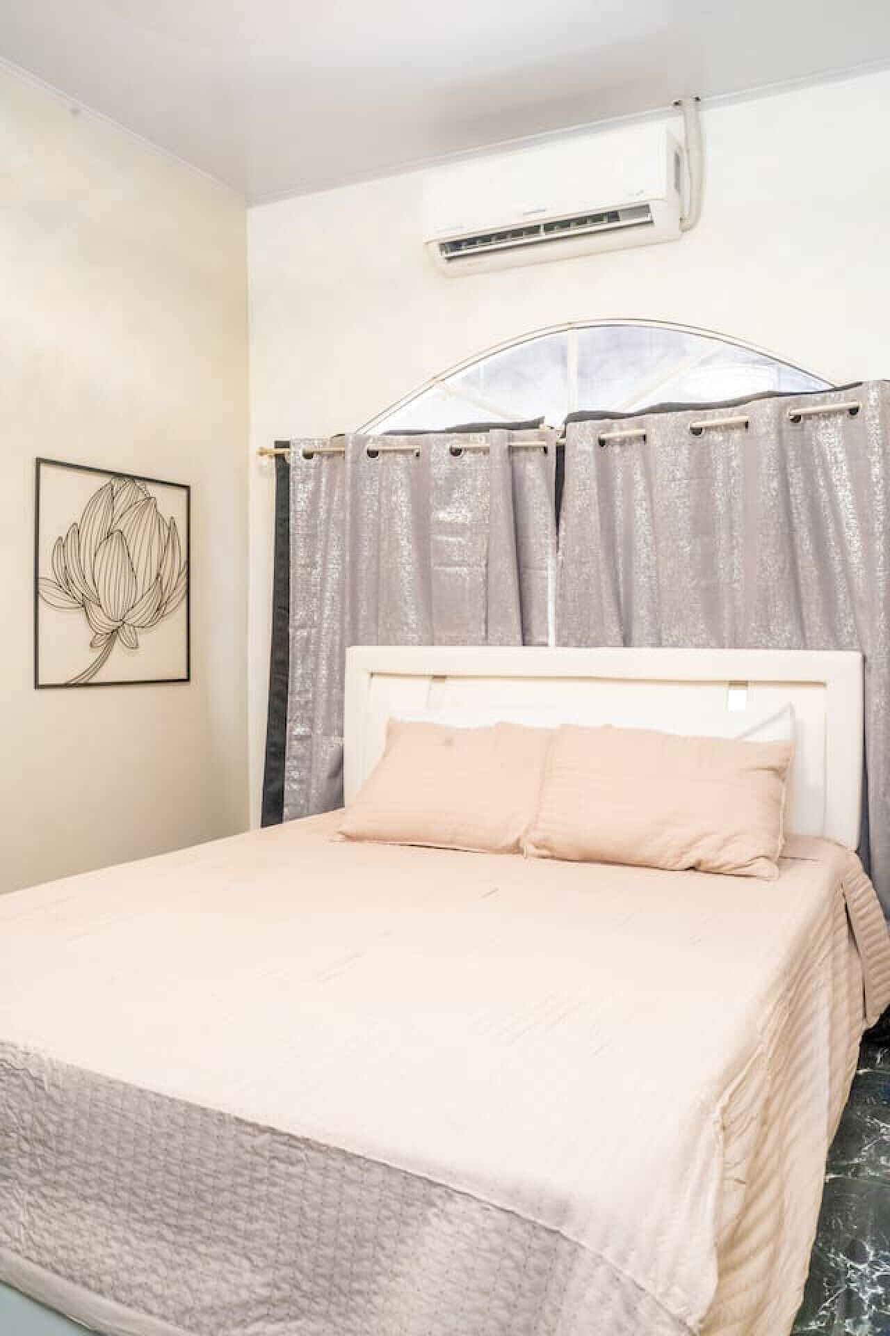 Un dormitorio con cama, cabecera, equipo de aire acondicionado, ventana con cortinas e imagen enmarcada en la pared.