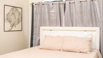 Un dormitorio con cama, cabecera, equipo de aire acondicionado, ventana con cortinas e imagen enmarcada en la pared.