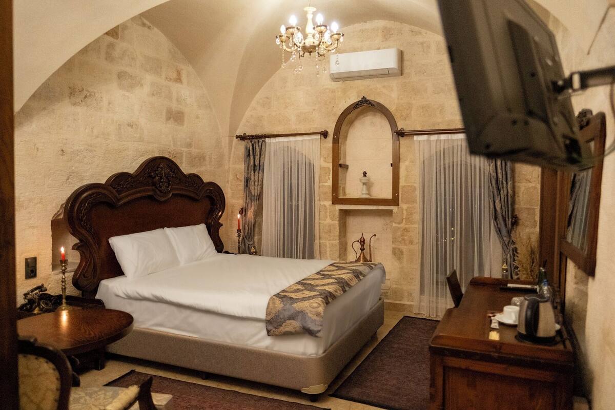 A bedroom with a large bed, a chandelier, a wooden headboard, a small table with a lamp, and a mirror.