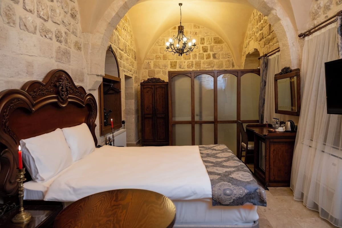 A bedroom with a large bed, wooden headboard, a chandelier, stone walls, and a wooden wardrobe.
