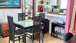 Un coin repas avec une table à plateau en verre, des chaises en bois, un vase rouge et, au fond, une cuisine.