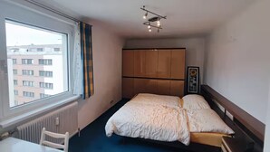 A hotel room with a bed, a desk, a chair, a radiator, a window, and a view of buildings outside. - Charming Vintage Vienna Getaway  67m² with A/C (Wien)