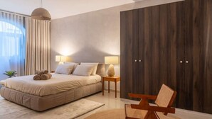 A modern bedroom with a large bed, two bedside lamps, a wooden wardrobe, and a woven chair.