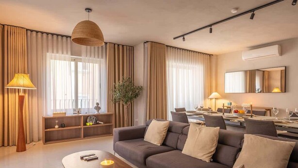 A modern hotel room with a dark grey sofa, a wooden coffee table, and a large window with sheer curtains.