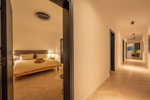 A modern hotel room with a large bed, bedside tables, and a hallway leading to other rooms.