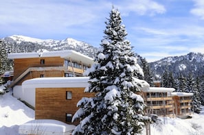 A modern ski resort building with multiple floors, wooden exterior, and a snow-covered mountain backdrop.