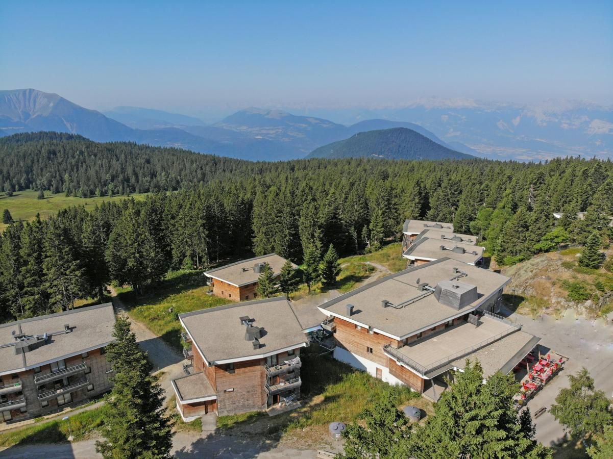 A cluster of modern buildings with flat roofs, surrounded by dense forest and mountains in the background.