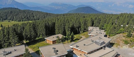 A cluster of modern buildings with flat roofs, surrounded by dense forest and mountains in the background.