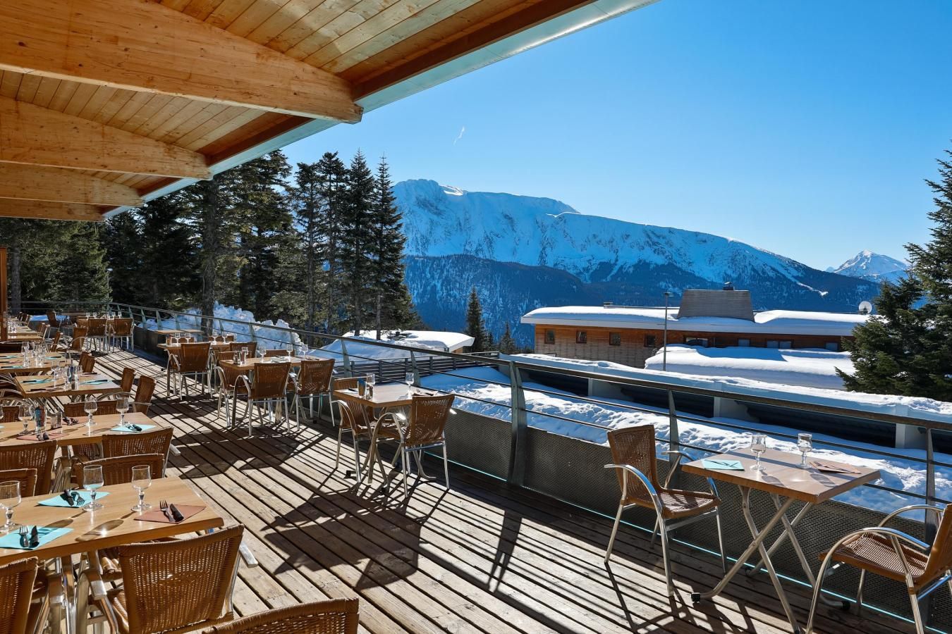 An outdoor dining area with wooden tables and chairs, overlooking a snowy mountain landscape and a building with a snow-covered roof.