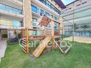 A playground with a wooden slide and climbing structure, surrounded by a well-maintained lawn and a building with multiple balconies in the background. - Condomínio Bela Vista Porto de Galinhas (Ipojuca)