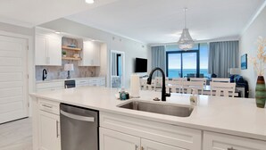 A modern kitchen with white cabinetry, a stainless steel sink, and a dining area with a view. - Premium Condo Beachfront Lazy River-Water slide-Splash Pad-Walk to Restaurants (Gulf Shores)