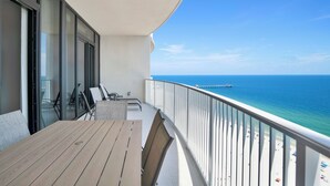 A balcony with a wooden table and chairs, a glass door, and a view of the ocean and pier. - Premium Condo Beachfront Lazy River-Water slide-Splash Pad-Walk to Restaurants (Gulf Shores)