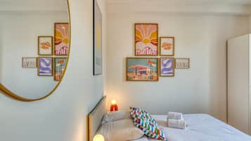 Een net opgemaakt bed met witte lakens en twee handdoeken, een kleurrijk geruit kussen, en een muur met lijstwerken, waaronder de woorden 'moment' en 'voyage'.