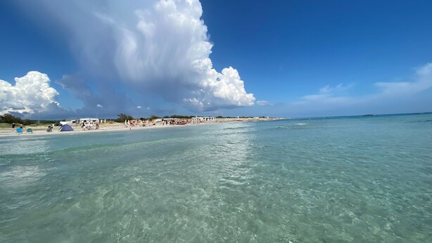Ein Strand mit klarem türkisblauem Wasser, einem sandigen Ufer und einer großen Wolke am Himmel.