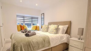 A neatly arranged bedroom with a bed, pillows, a bedside table, and a window with blinds. - Bubbles & Bliss - Hot Tub/Garden | Pass the Keys (Rye)