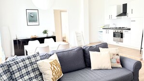 Ein modernes Wohnzimmer mit grauem Sofa, weißen Stühlen, einem Esstisch und einer Küchenzeile mit weißen Schränken und Geräten.