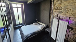 A modern bedroom with a curved bed, a small bedside table, a window with curtains, and a stone wall. - A charming gite (Sainte-Luce-sur-Loire)