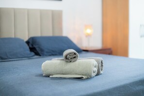 Una cama bien hecha con una toalla doblada, un cabecero y una lámpara de noche.