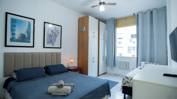 Un dormitorio con cama, escritorio, ventilador de techo y dos cuadros en la pared.