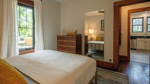 Ein Schlafzimmer mit Bett, Kommode, einem Fenster mit Vorhängen, einem Spiegel und Blick auf eine Küche.