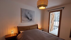 Ein Schlafzimmer mit einem Bett, einem Nachttisch mit Lampe, einem Wandbild und einem Fenster mit Blick auf eine verschneite Landschaft.