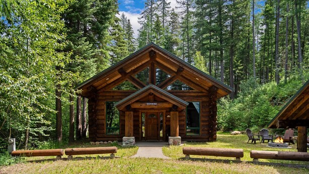 A log cabin with a central entrance, surrounded by a natural forest setting.