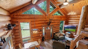 A log cabin interior with a dining area, a fireplace, and a large window overlooking a scenic landscape.