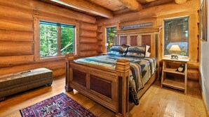 A log cabin bedroom with a wooden bed, a bench, and a window with a view of trees.