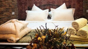 Una cama bien tendida con sábanas y almohadas blancas, un arreglo floral sobre la mesita de noche y una pared de ladrillo al fondo.