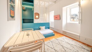 Une pièce de taille modeste comprenant un lit, une petite table avec des chaises, un meuble de rangement bleu, une fenêtre donnant sur la ville et un tableau encadré accroché au mur.