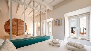 A modern hotel room with a large bed, a round mirror, a view of the street, and a window with potted plants. - GuestReady - Esperança do Cardal 2D (Lisbon)