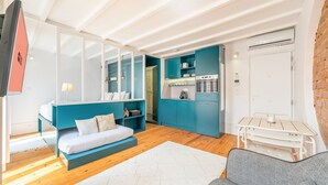 Ein kompakter Wohnbereich mit Sofa, einem kleinen Esstisch, einer Küchenzeile und einem durch eine Glastür einsehbaren Badezimmer.