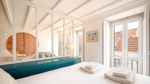 Ein Hotelzimmer mit einem großen Bett, einer blauen Kommode, einem Spiegel, einem Fenster mit Blick auf Gebäude und einem runden Spiegel an der Wand.