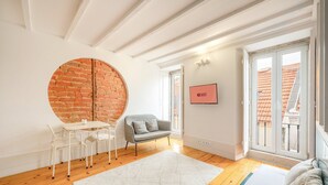 Ein modernes Zimmer mit Backsteinwand, einem runden Fenster, einem weißen Tisch mit Stühlen, einem grauen Sofa und einem Fernseher an der Wand.