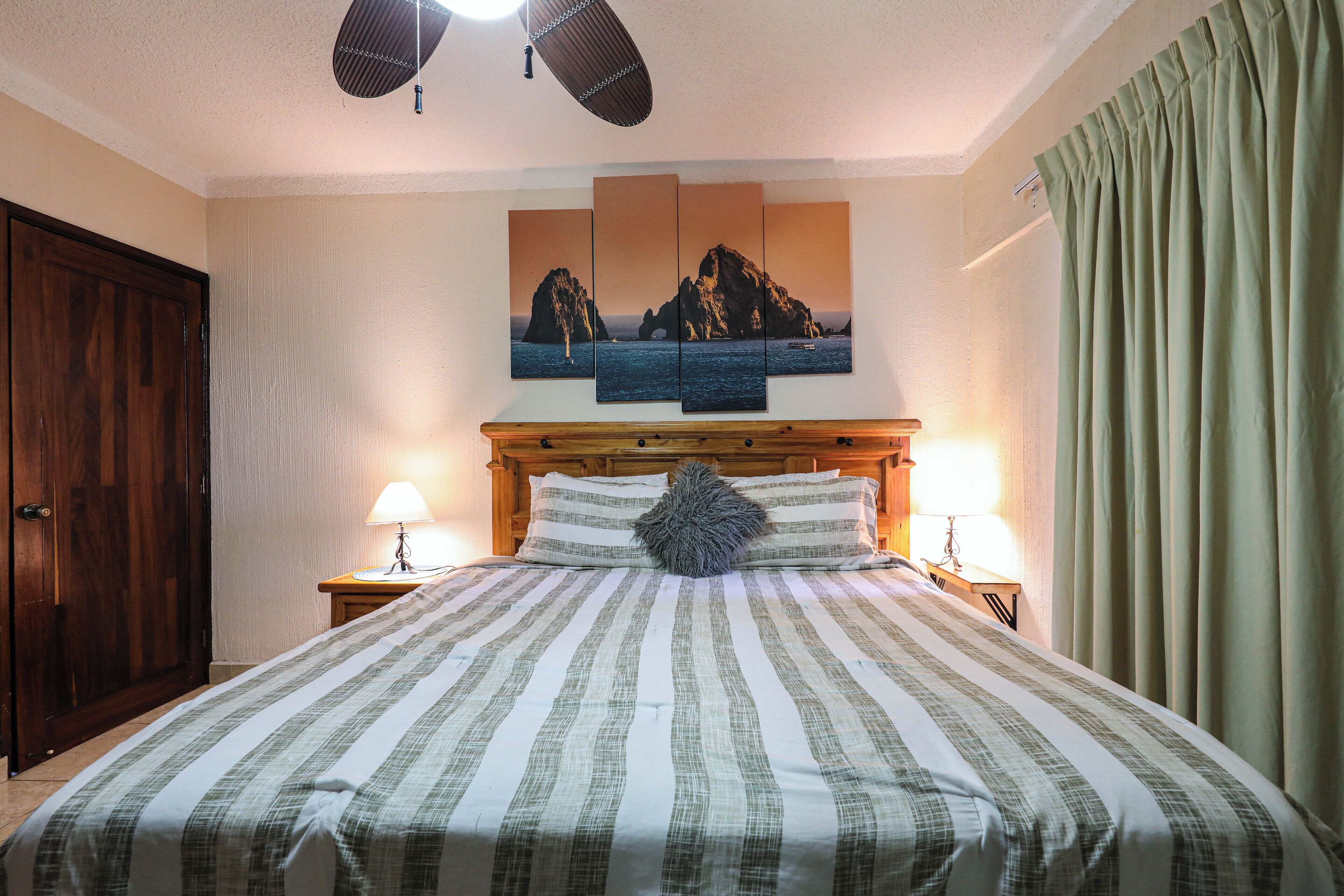 A hotel room with a large bed, a wooden headboard, a painting of a coastal scene, two bedside lamps, and a green curtain.
