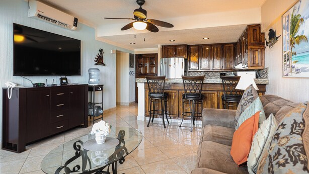 A spacious living area with a large flat-screen TV, a ceiling fan, and a kitchen with wooden cabinets and a bar area.
