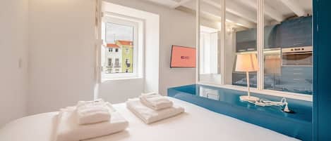 A hotel room with a bed, a window with a view of buildings, a mirror, a TV, and a blue counter.