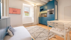 Habitación de hotel pequeña con cama, una mesita, kitchenette y ventana con vistas a edificios.