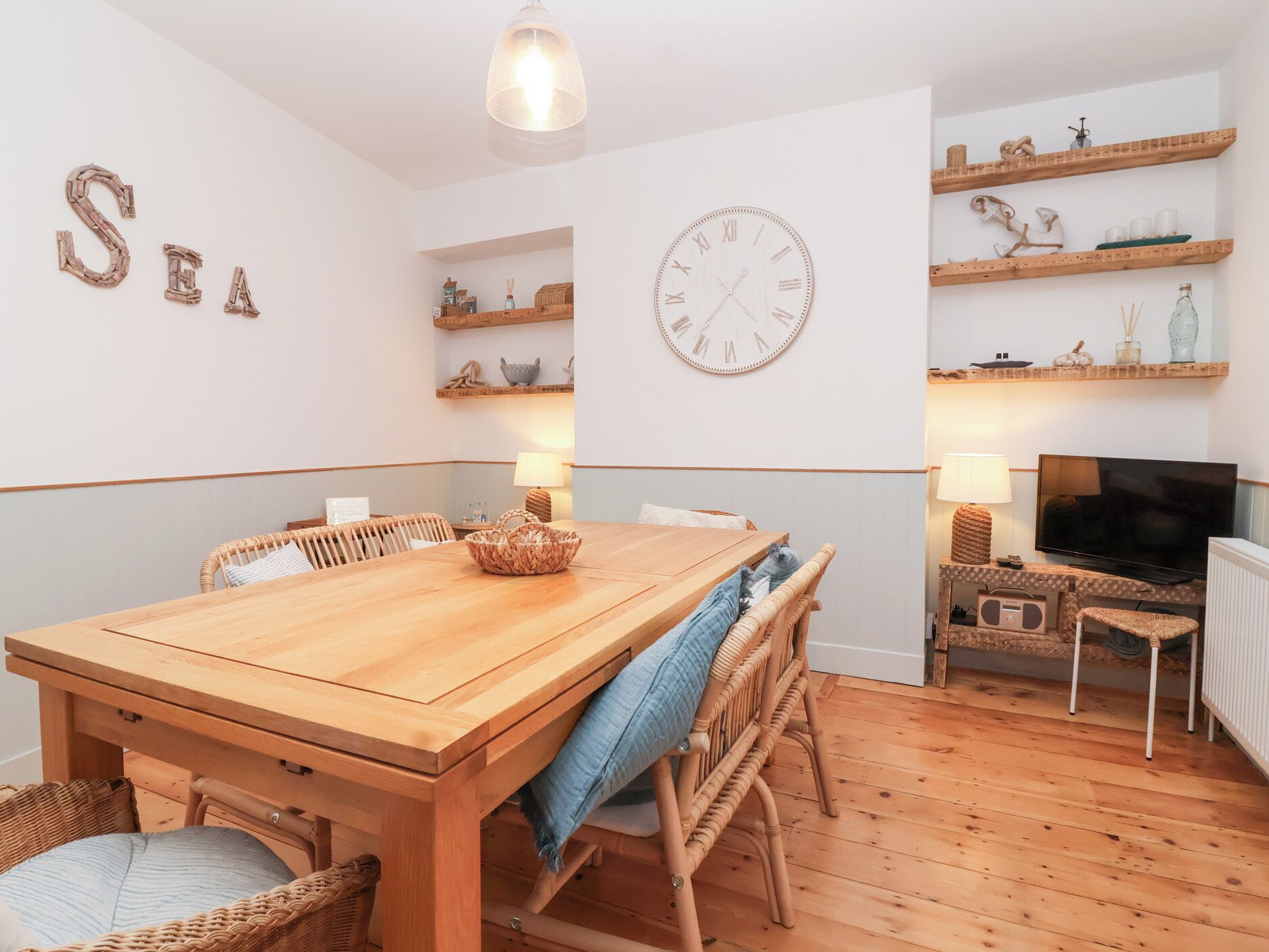 Um quarto aconchegante com uma mesa de jantar de madeira, cadeiras de vime, televisão e prateleiras com objetos decorativos.