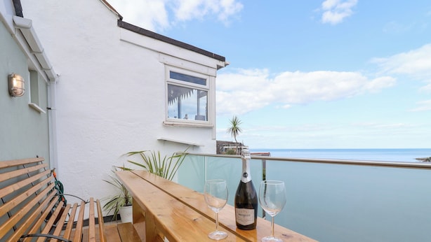 Banco y mesa de madera con una botella de Prosecco y dos copas en un balcón con vistas al mar.