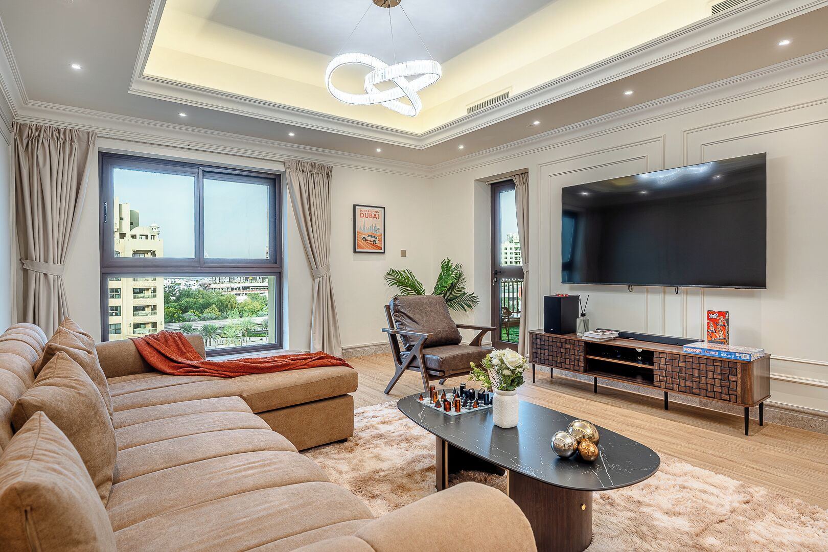 Un salon moderne avec un canapé d’angle, une table basse, un téléviseur et une grande fenêtre donnant sur la ville.