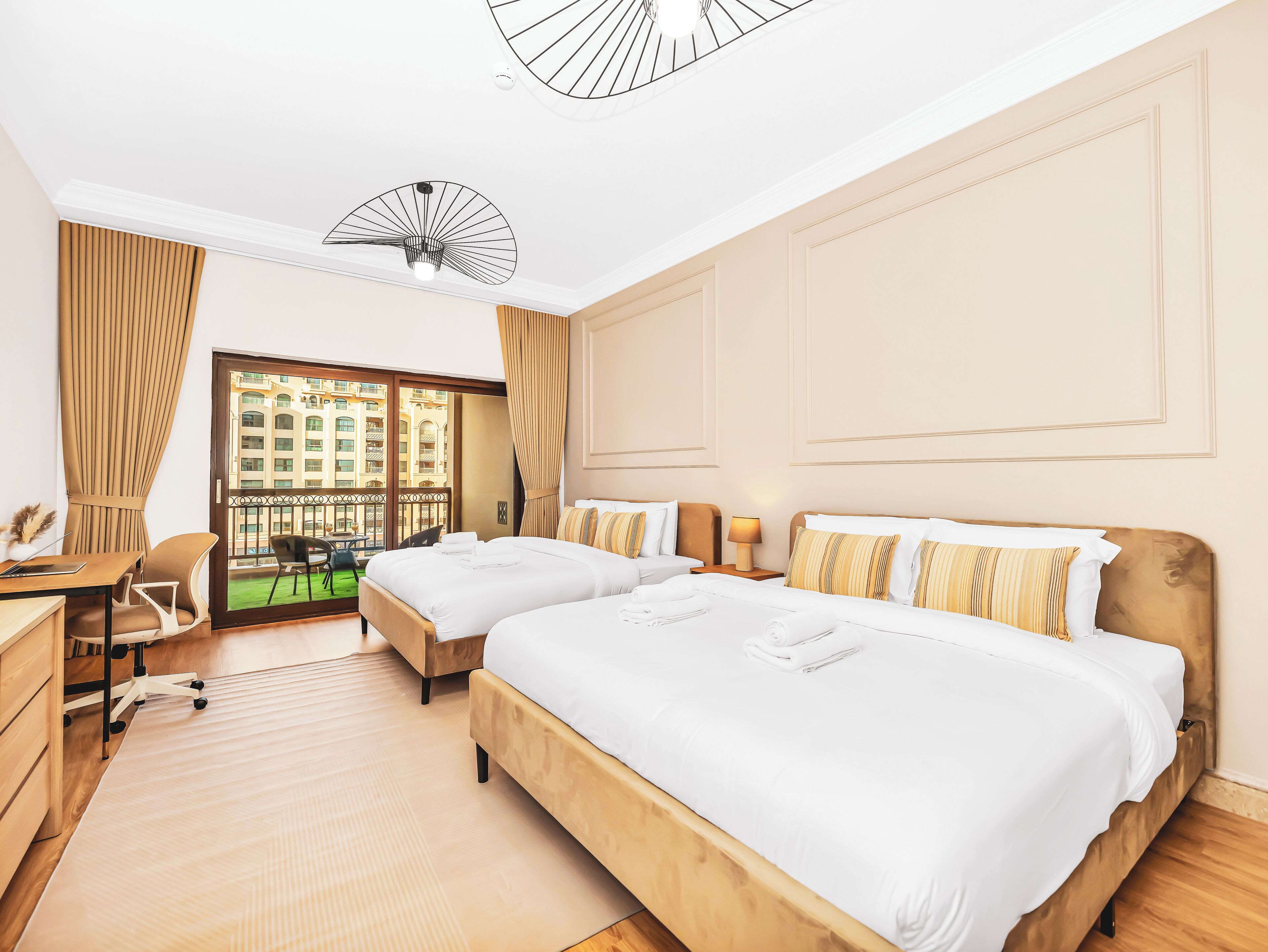Ein Hotelzimmer mit zwei Betten, einem Schreibtisch, einem Stuhl und Blick auf ein Gebäude durch eine Glastür.