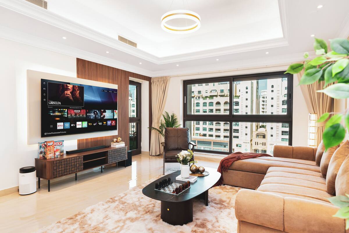 Een moderne woonkamer met een grote flatscreen televisie, een comfortabele bank, een salontafel en een uitzicht op een stadsbeeld door grote ramen.
